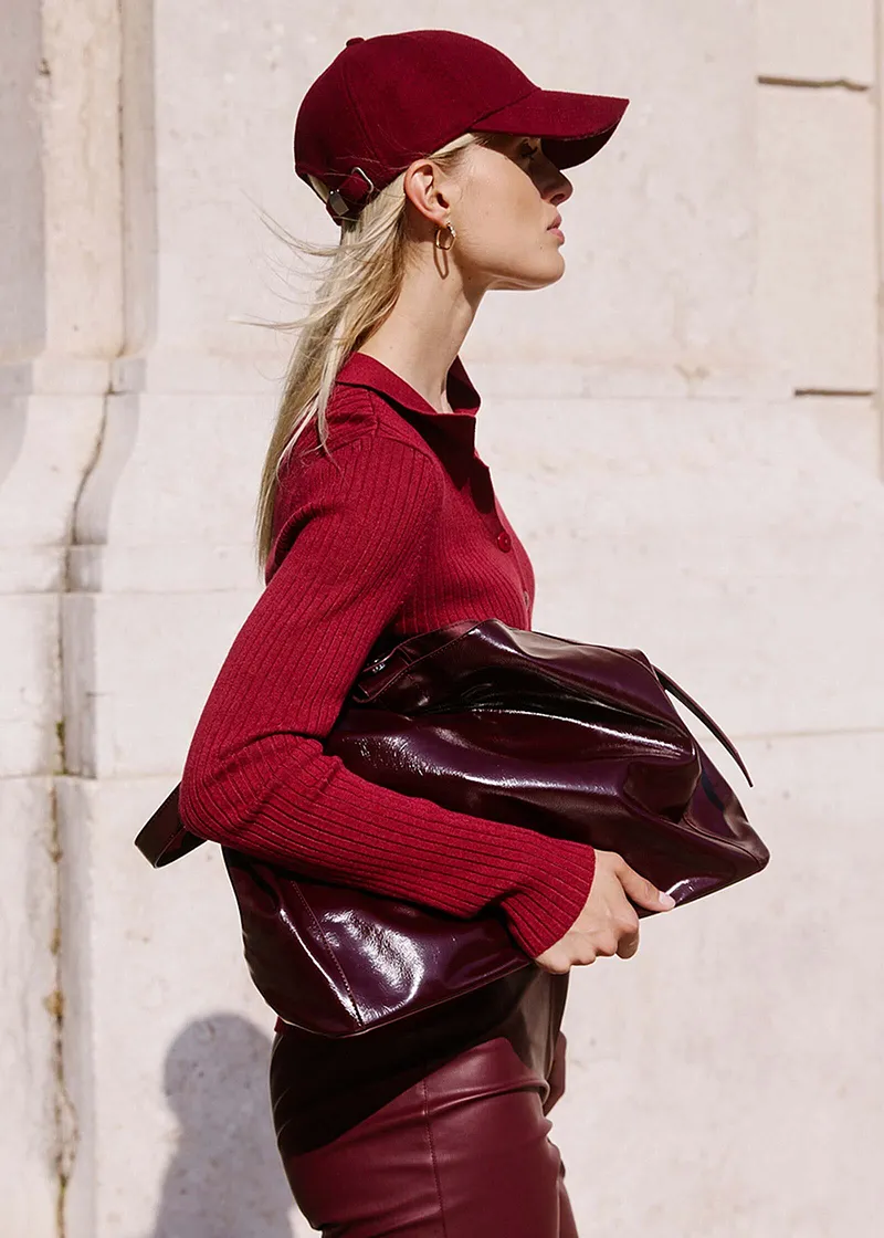 Elegante bordeauxrode look: gebreide trui, leren broek, baseball pet en een glanzende handtas.