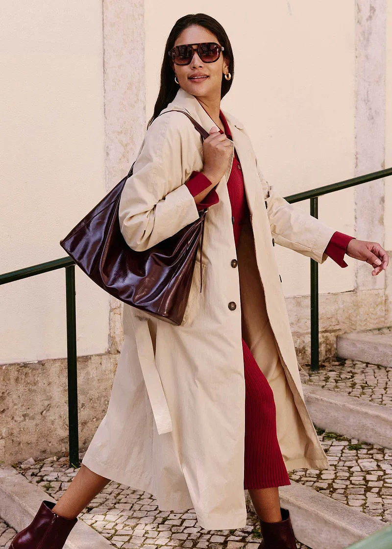 Elegante beige trenchcoat over rode jurk, met bruine laarsjes en grote tas.
