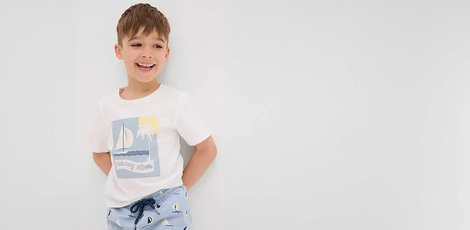 Fröhliches Kind in weißem T-Shirt mit Segelboot-Motiv und hellblauer Hose mit Segelboot-Muster.