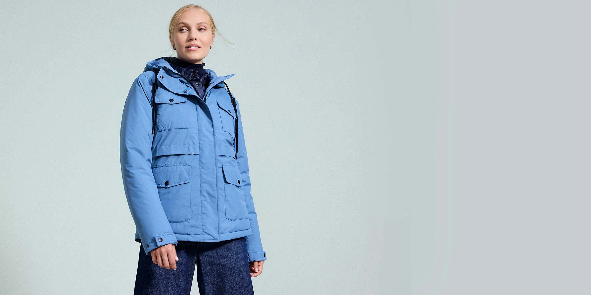 Lichtblauwe winterjas met capuchon en meerdere zakken, gecombineerd met donkere sjaal en jeans.