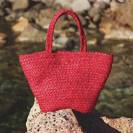 Felrode rieten handtas rust op rotsen bij het water, perfect voor zomerse stranduitstapjes.