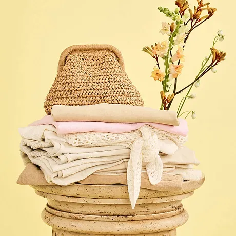 Natuurlijke tinten: rieten tas, delicate dekens en een wit kledingstuk op een houten zuil.