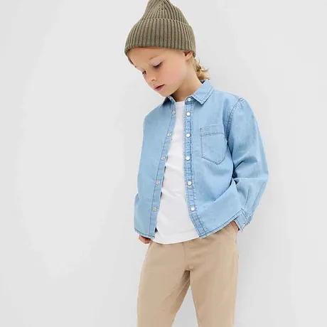 Lichtblauw spijkerhemd over wit T-shirt, beige broek en groene gebreide muts: een casual kinderoutfit.