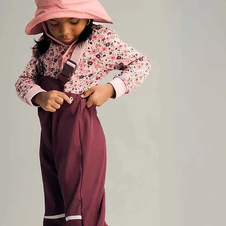 Vrolijke meisjeslook: gebloemde roze trui, bordeauxkleurige regenbroek en een pastelkleurige zonnenhoed.