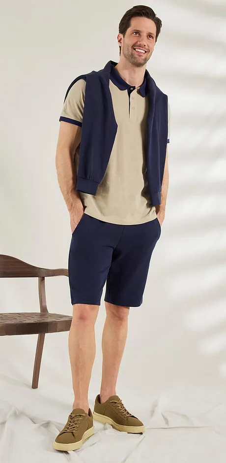Casual zomerlook: beige poloshirt, donkerblauwe trui en shorts, met bruine sneakers. Relaxte style.