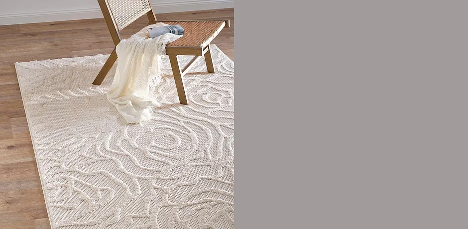 Cremefarbene Decke liegt lässig über Rattanstuhl auf elegantem Teppich mit geschwungenen Mustern.