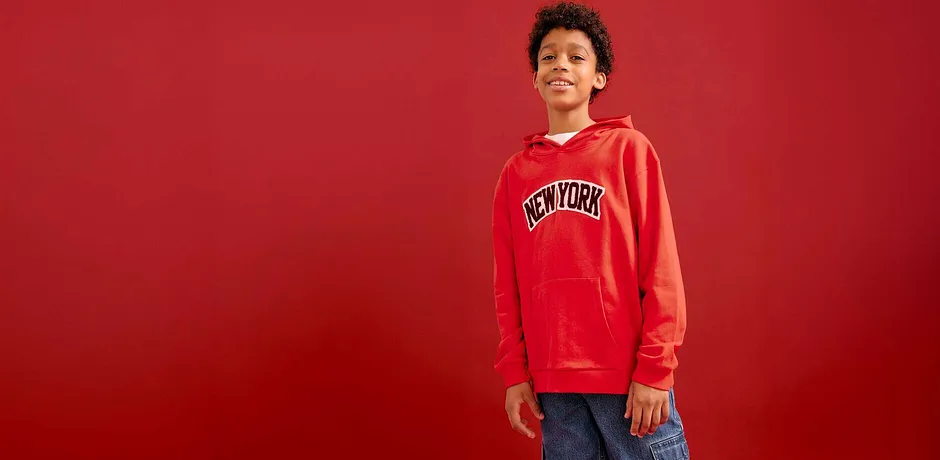 Roter Kapuzenpullover mit Aufdruck, blaue Jeans. Lässiger Streetwear-Look vor rotem Hintergrund.
